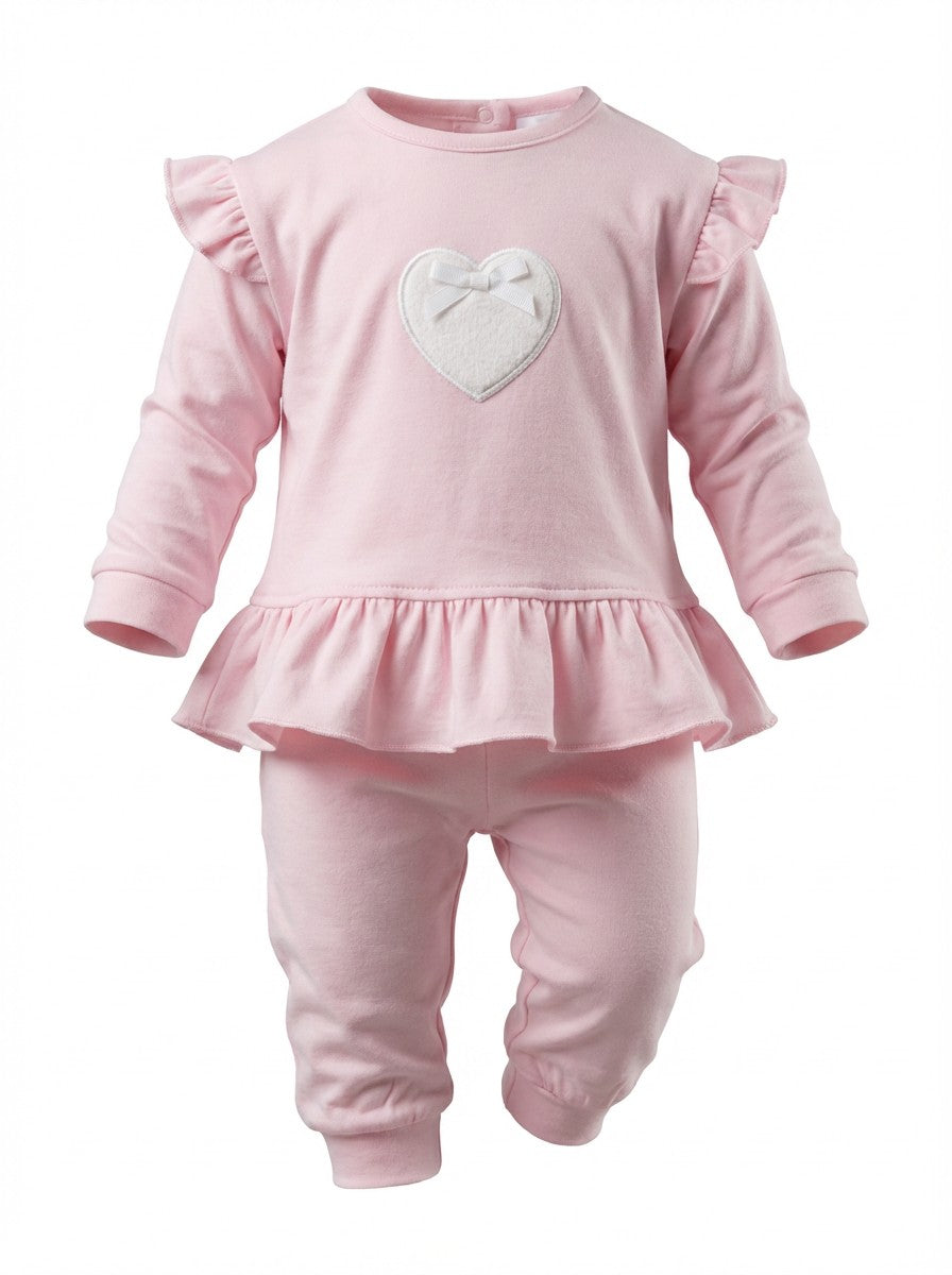 A baby girl's two-piece outfit in light pink, featuring a long-sleeve top with ruffle details on the shoulders and a ruffled hem, paired with matching pants. The top has a white applique heart on the chest with a small bow detail.